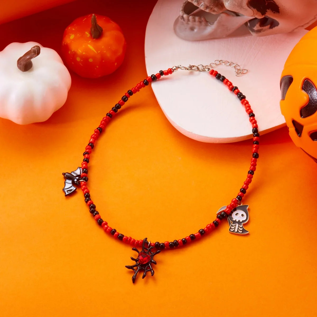 Collier Halloween en perles rouges et noires avec pendentifs chauve-souris, fantôme et araignée, sur fond orange avec citrouilles