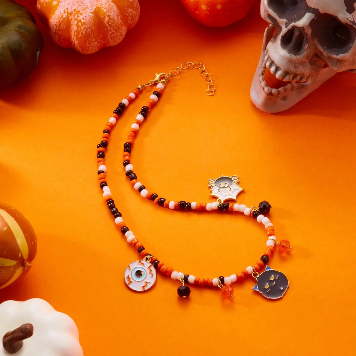 Collier Halloween en perles orange, blanches et noires avec pendentifs œil, chat et toile d'araignée, sur fond orange avec crâne et citrouilles