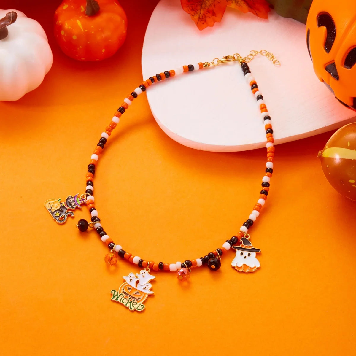 Collier Halloween avec pendentifs fantôme, citrouille et chauve-souris, présenté à plat sur fond orange avec citrouilles