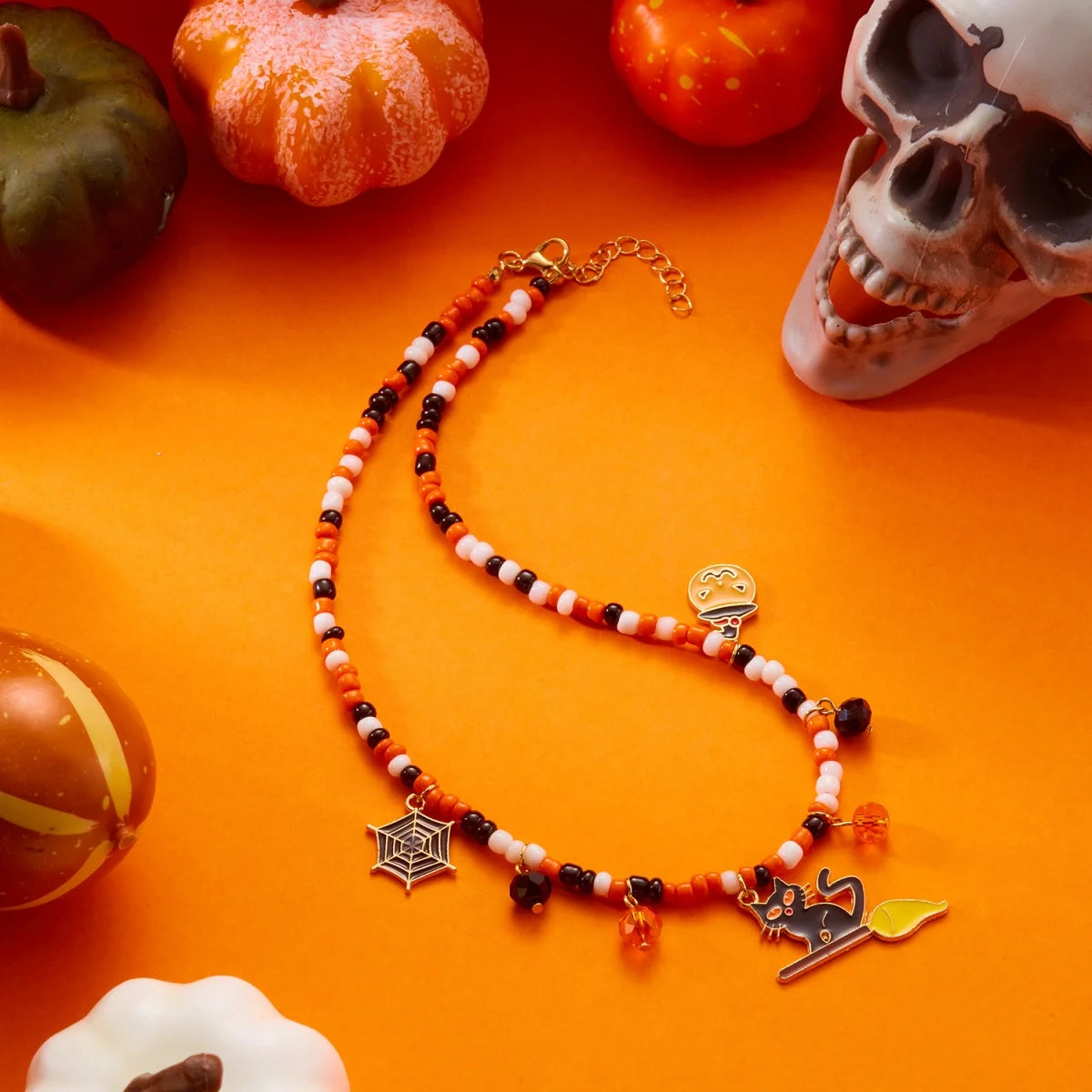 Collier Halloween en perles orange, blanches et noires avec pendentifs toile d'araignée, fantôme et chat noir, sur fond orange avec citrouilles et crâne
