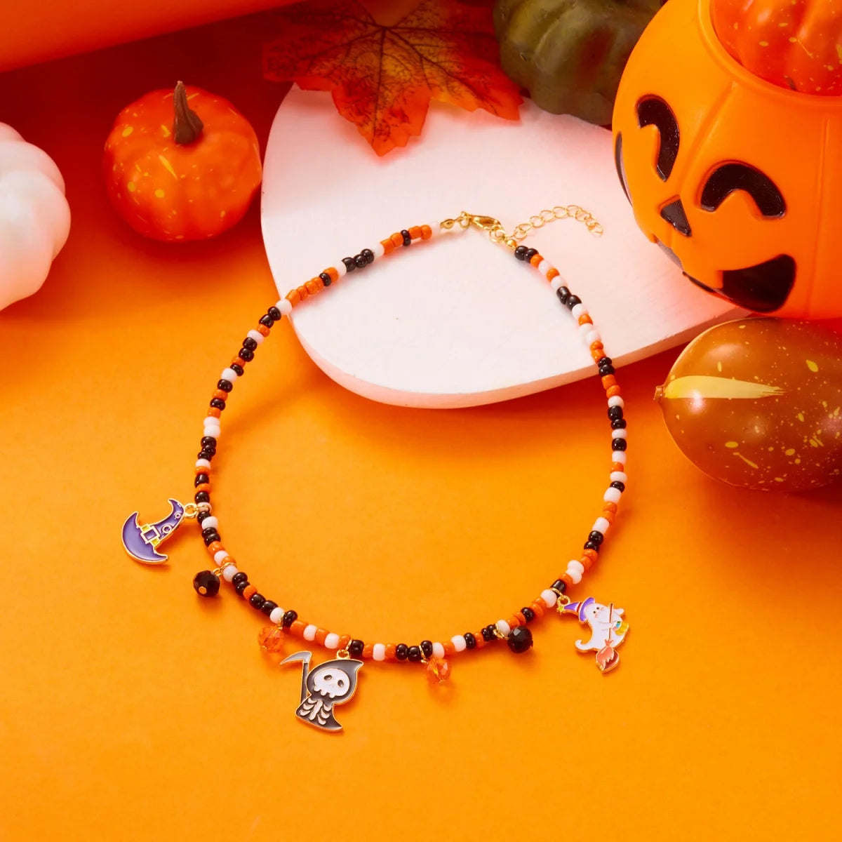 Collier Halloween en perles orange, blanches et noires avec petits pendentifs lune, fantôme et chat, sur fond orange avec citrouilles